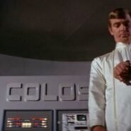 Homme en combinaison blanche devant un ordinateur nommé Colossus