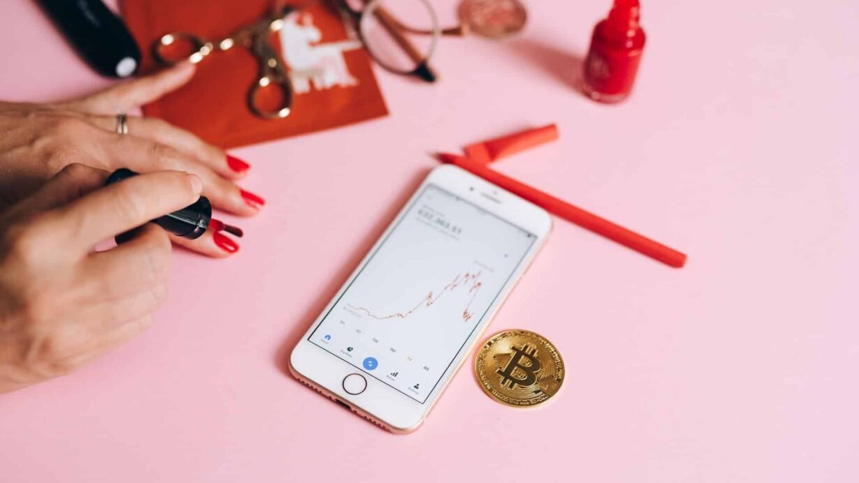 Main avec vernis à ongles rouge, téléphone affichant un graphique et pièce de bitcoin