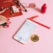 Main avec vernis à ongles rouge, téléphone affichant un graphique et pièce de bitcoin