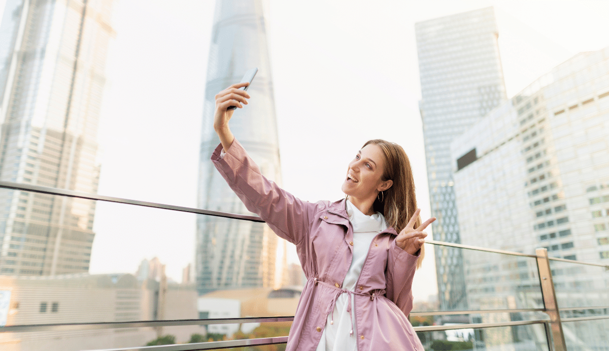 Femme souriante en veste rose prenant un selfie devant des gratte-ciel