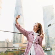 Femme souriante en veste rose prenant un selfie devant des gratte-ciel