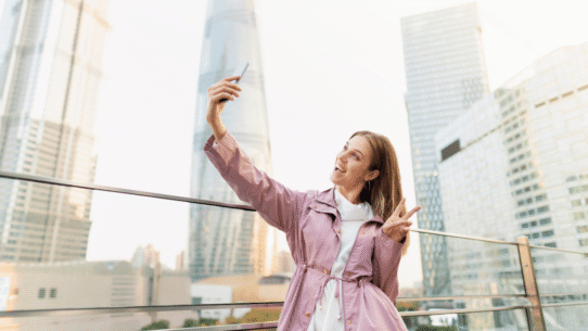 Femme souriante en veste rose prenant un selfie devant des gratte-ciel