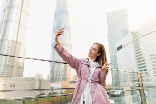 Femme souriante en veste rose prenant un selfie devant des gratte-ciel