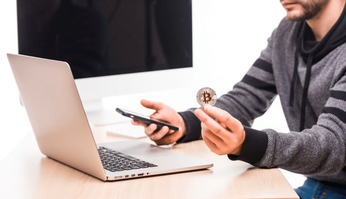 Personne tenant un Bitcoin et un smartphone devant un ordinateur