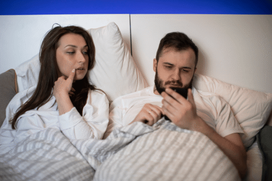 Un couple au lit, l'homme regarde un smartphone